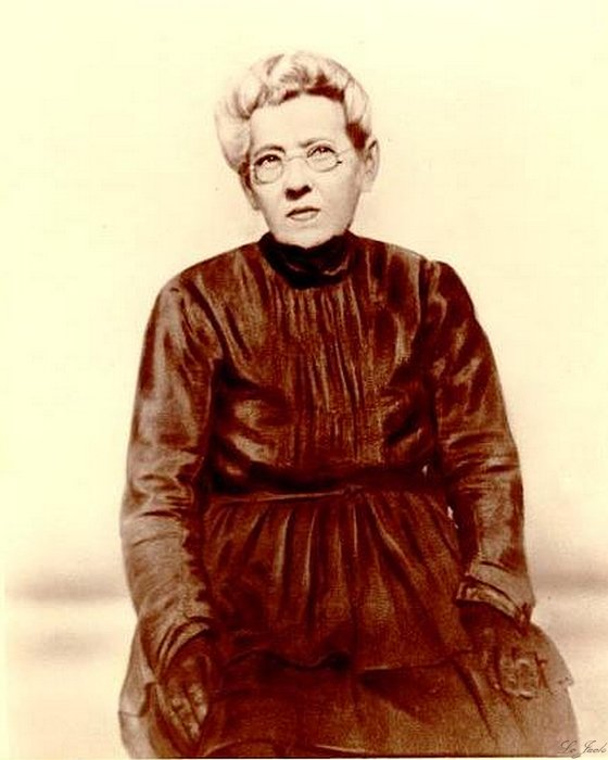 Sepia studio portrait of Kitty (Katie) Prater, seated, wearing round glasses and a dark high-necked dress