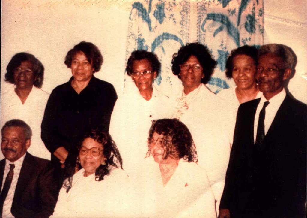 Faded color photo of nine of Warren Prater's children, two men in suits and seven women, posed together indoors in front of a patterned curtain